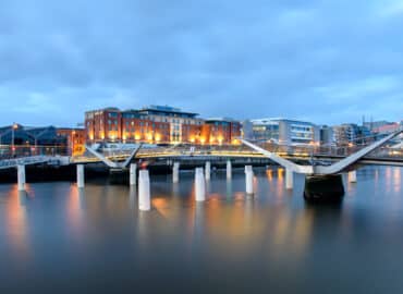 A white, steel pedestrian bridge with conical white supporting posts. Photographed in twilight, lit from behind with amber-coloured lights.