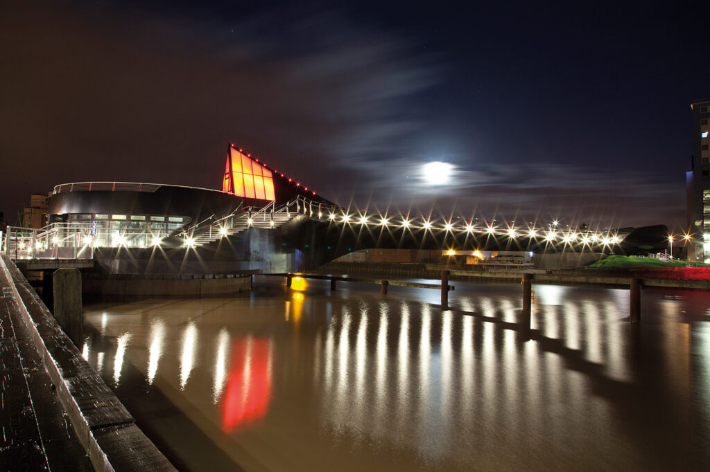 Scale Lane Bridge photographed from the side of the River Hull at night-time, against a black sky. The bright white moon is partially cloud-covered above the bridge. The bridge is illuminated with orange and white lights, reflected in the light-brown water of the River Hull.