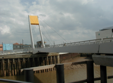 The Millennium Bridge photographed from the side of the river Hull against a blue, cloudy sky. The bridge is 'L' shaped and painted white with a bright yellow counterweight at its top edge.