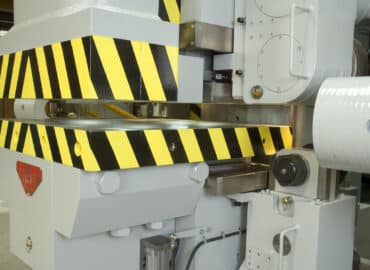 Close-up view of a light grey metal frame bending machine with striking yellow and black safety guard markings.