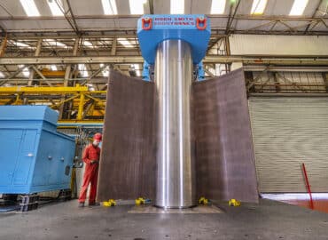 An engineer wearing red overalls stands beside a vertical plate bending machine loaded with a curved metal sheet.