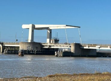A white, dutch-style road bridge in its closed position to allow traffic to pass over the River yare. Pictured from a distance on a clear, sunny day against a blue sky.