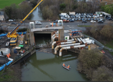 A drone photograph taken above the Woodhouse Mill Regulator. A series of large white pipes run from the riverside into the dark green river water. A yellow crane can be seen on the left of the picture and around 30 cars and vehicles parked nearby to the right of the picture. Workmen in orange hi vis are in a small boat in the river, and on either side of the regulator.