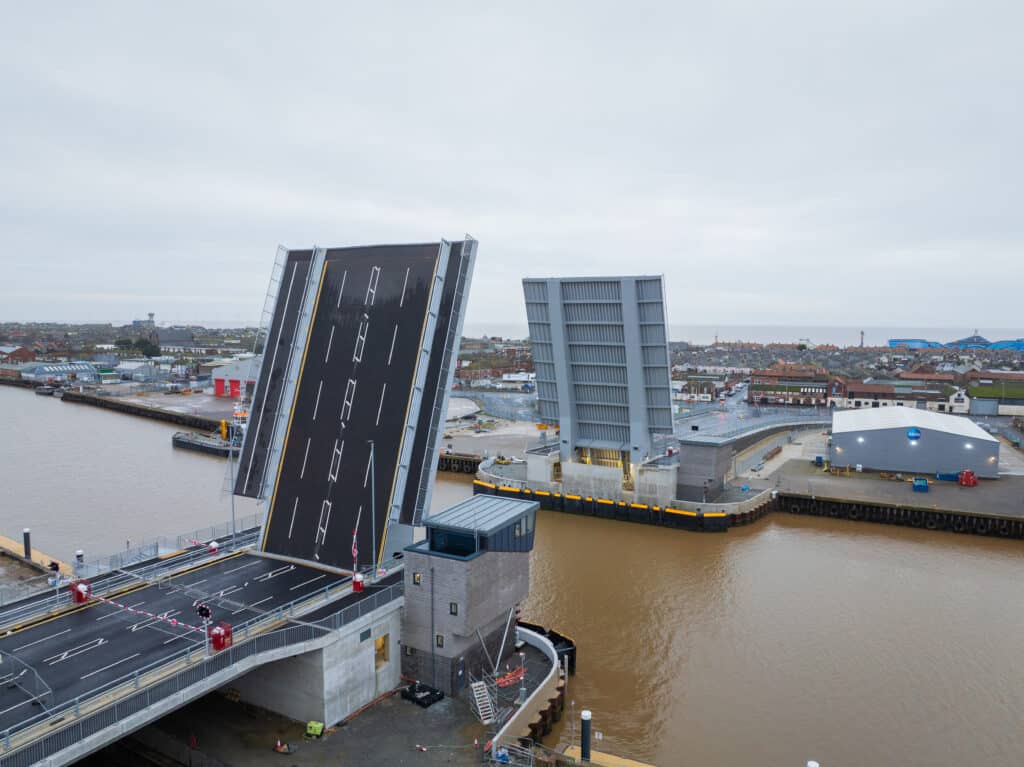 Herring Bridge pictured from above on a clear day, under a white/greay cloudy sky. The River Yare looks smooth and brown, and the bridge's bascule leaves are raised up so that cars cannot pass across the bridge, but large boats could pass by underneath.