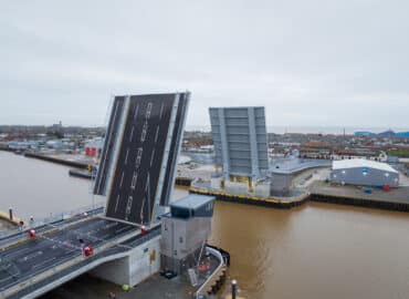 Herring Bridge pictured from above on a clear day, under a white/greay cloudy sky. The River Yare looks smooth and brown, and the bridge's bascule leaves are raised up so that cars cannot pass across the bridge, but large boats could pass by underneath.