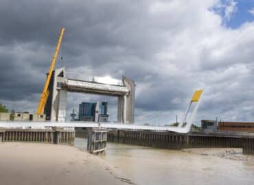 Hull's Millennium Bridge photographed from the bank of the River Hull. The bridge is white and forms an 'L' shape with a striking yellow counter-weight on its elevated side. The skiy is blue and full of white/grey clouds. The river is light-brown and the Hull Tidal Barrier can be seen in the distance, behind the bridge.