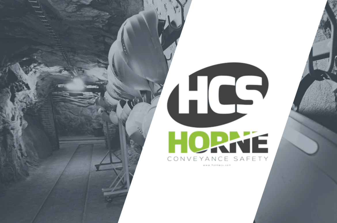 Hard hats hang on a rail in an underground tunnel with rough rocky walls and a wood floor