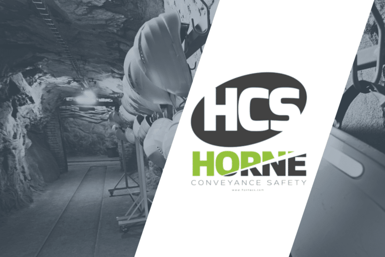 Hard hats hang on a rail in an underground tunnel with rough rocky walls and a wood floor
