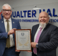 Gifford Brown MD of Qualter Hall wears a navy blue suit and stands beside Ivan Tibble, Environment Health and Safety Manager at Qualter Hall wearing a grey suit. They stand infront of a large Qualter Hall logo, smile and hold a RoSPA Presidents Award certificate in a brown wooden frame