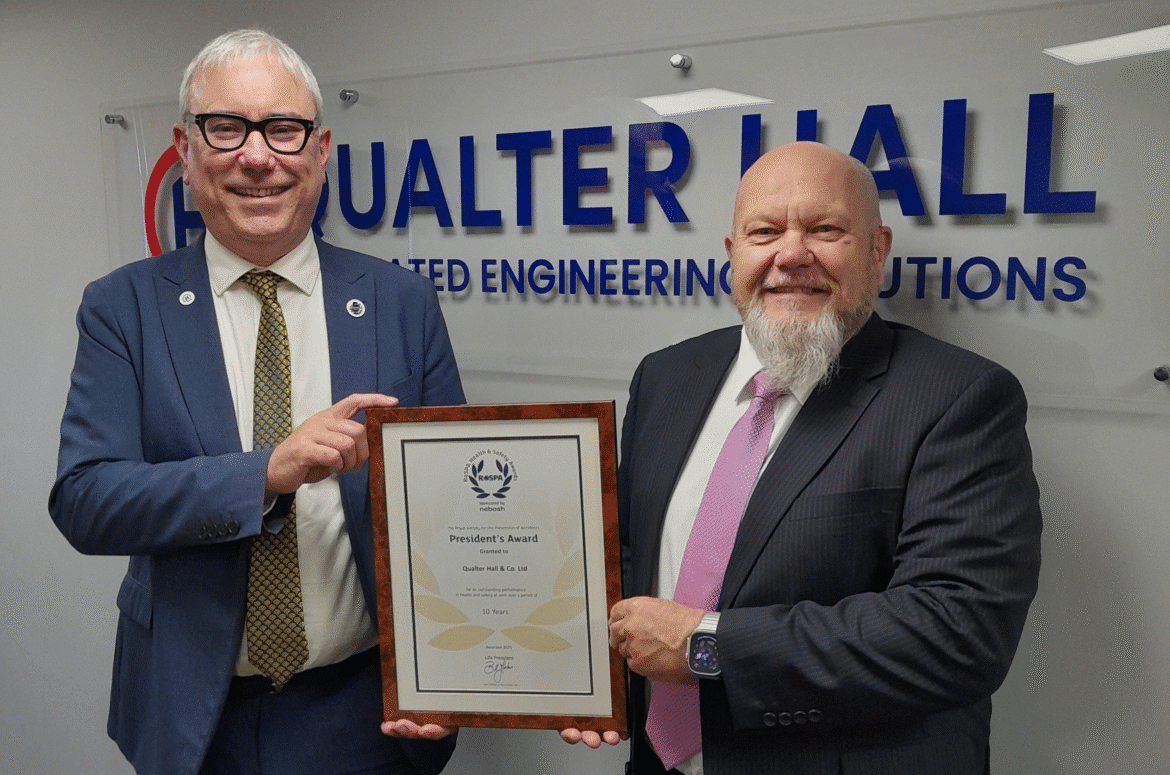 Gifford Brown MD of Qualter Hall wears a navy blue suit and stands beside Ivan Tibble, Environment Health and Safety Manager at Qualter Hall wearing a grey suit. They stand infront of a large Qualter Hall logo, smile and hold a RoSPA Presidents Award certificate in a brown wooden frame