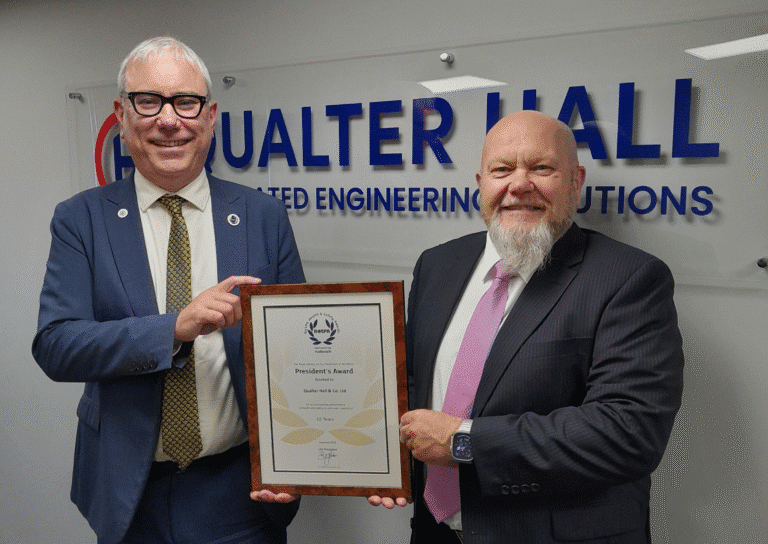 Gifford Brown MD of Qualter Hall wears a navy blue suit and stands beside Ivan Tibble, Environment Health and Safety Manager at Qualter Hall wearing a grey suit. They stand infront of a large Qualter Hall logo, smile and hold a RoSPA Presidents Award certificate in a brown wooden frame