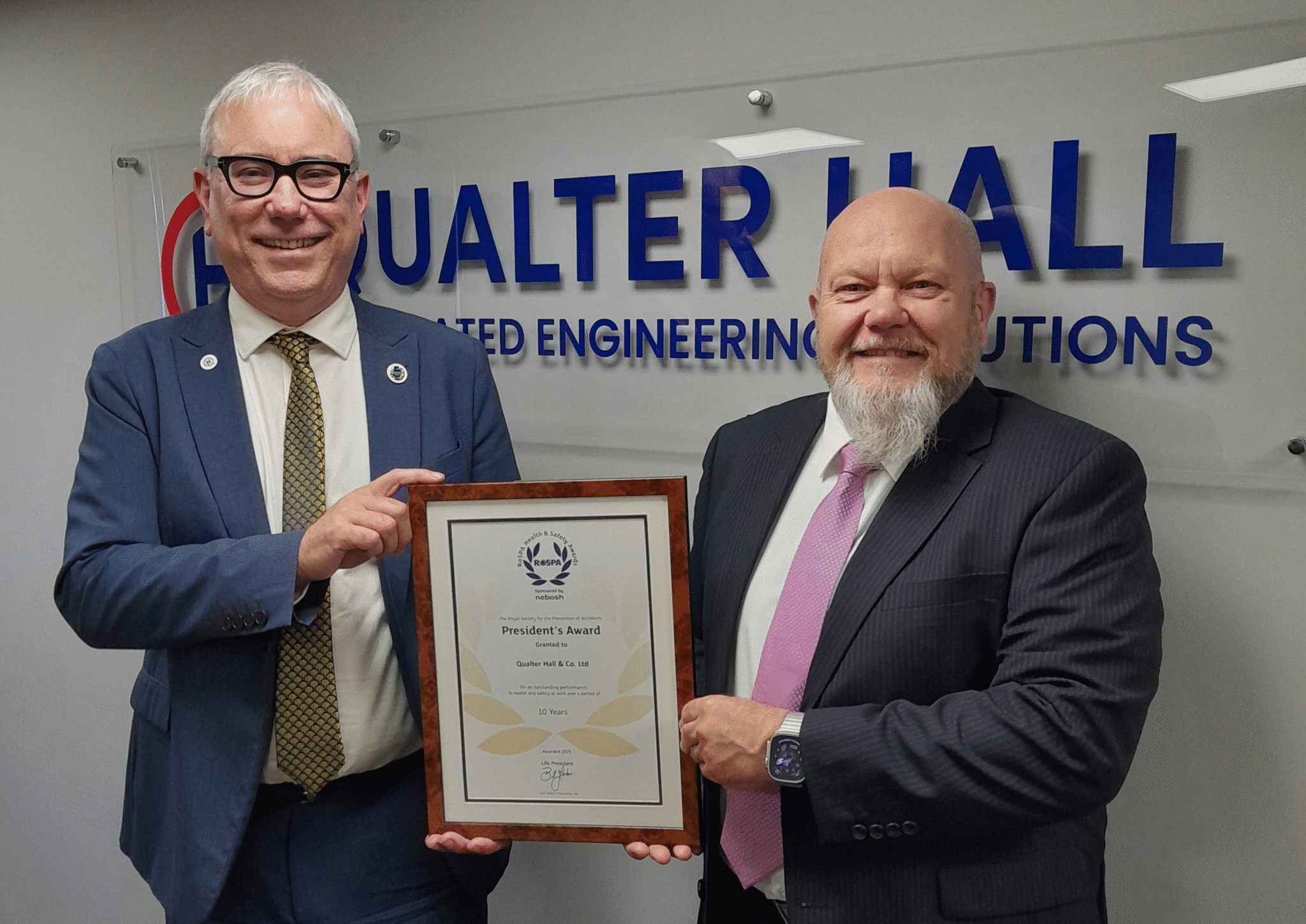Gifford Brown MD of Qualter Hall wears a navy blue suit and stands beside Ivan Tibble, Environment Health and Safety Manager at Qualter Hall wearing a grey suit. They stand infront of a large Qualter Hall logo, smile and hold a RoSPA Presidents Award certificate in a brown wooden frame