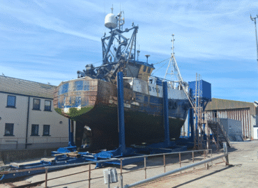 A royal blue painted steel cradle sits at the top of a harbour slipway. A boat is held in the cradle's vertical bilge arms, which stand along the cradle on either side of the boat. The sky is cloudless and blue, and and we can see the grey, concrete slipway in the foreground of the image, a grey, pipework metal fence and cream-coloured buildings with dark rectangular windows at the slide of the slipway.