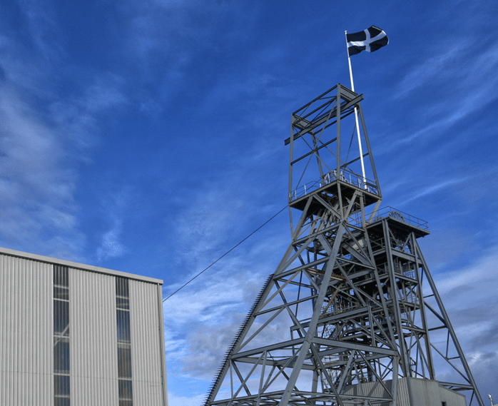 The grey steel headframe above the Cook's Kitchen Shaft at South Crofty Tin Mine, against a blue sky streaked with fluffy white clouds.