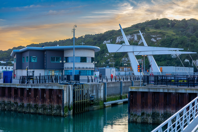 A bascule bridge with white, angular counterweights stands over a turquoise waterway at dusk. To the left is a modern, two-story harbor building with a curved glass facade, situated against a backdrop of steep green cliffs and a golden sunset sky.