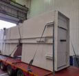 10T steel skip sitting on a flat bed lorry in Qualter Hall's Barnsley workshop, awaiting dispatch. The skip is pained a light beige and is strapped down on the red lorry trailer.