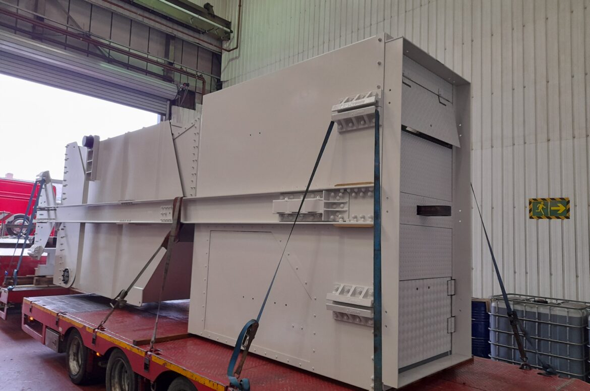 10T steel skip sitting on a flat bed lorry in Qualter Hall's Barnsley workshop, awaiting dispatch. The skip is pained a light beige and is strapped down on the red lorry trailer.
