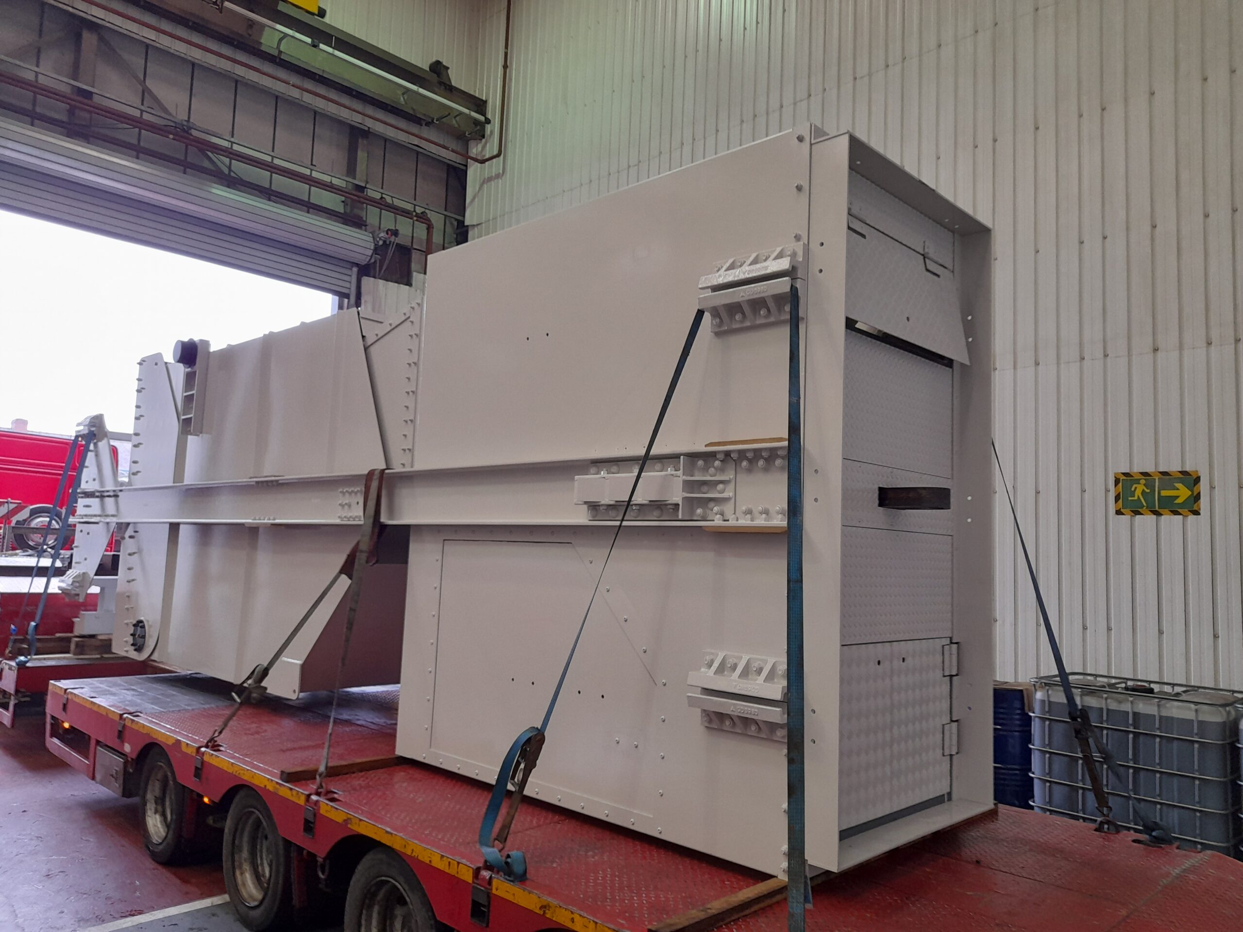 10T steel skip sitting on a flat bed lorry in Qualter Hall's Barnsley workshop, awaiting dispatch. The skip is pained a light beige and is strapped down on the red lorry trailer.
