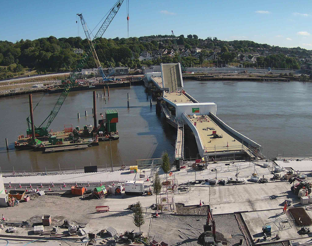 A view from above, of the bridge under construction. Two cranes are floating on barges alongside the bridge. The river water is calm and brown-coloured. The bridge deck is light brown with white edges. One bascule leaf is partially open and we can see a multitude of orange traffic cones on the bridge deck, and several people working in orange hi-vis PPE workwear and white hard hats. Trees and houses can be seen on the shore in the distance and the sky is bright blue with small white clouds.