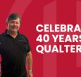 Carl smiles and wears his grey Qualter Hall workshop overalls with a black t-shirt and protective glasses. Andrew stands beside him, wearing a black branded Qualter Hall polo shirt.