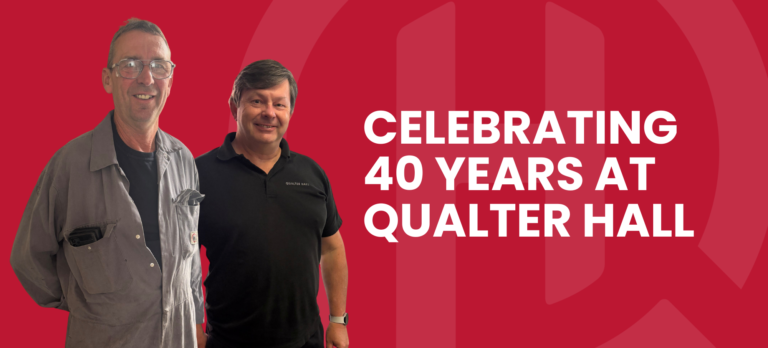 Carl smiles and wears his grey Qualter Hall workshop overalls with a black t-shirt and protective glasses. Andrew stands beside him, wearing a black branded Qualter Hall polo shirt.