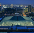 An aerial view of the British Museums' Great Consort Roof, set against a London skyline featuring landmarks such as the Millennium Wheel lit-up against a bright blue night sky.