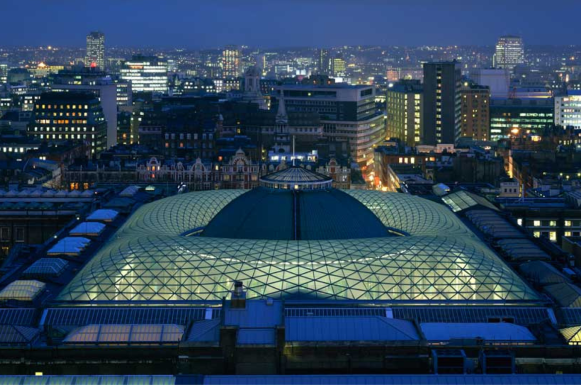 An aerial view of the British Museums' Great Consort Roof, set against a London skyline featuring landmarks such as the Millennium Wheel lit-up against a bright blue night sky.