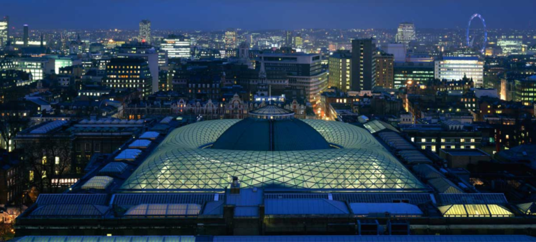 An aerial view of the British Museums' Great Consort Roof, set against a London skyline featuring landmarks such as the Millennium Wheel lit-up against a bright blue night sky.