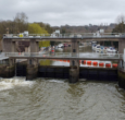 Allington Weir on the River Medway, Kent. Photographed from above showing trees on either side and a grey, cloudy sky. White water is rushing through the sluice gate and boats can be seen on the higher side of the river beyond, behind large orange bouys laid out across the weir.