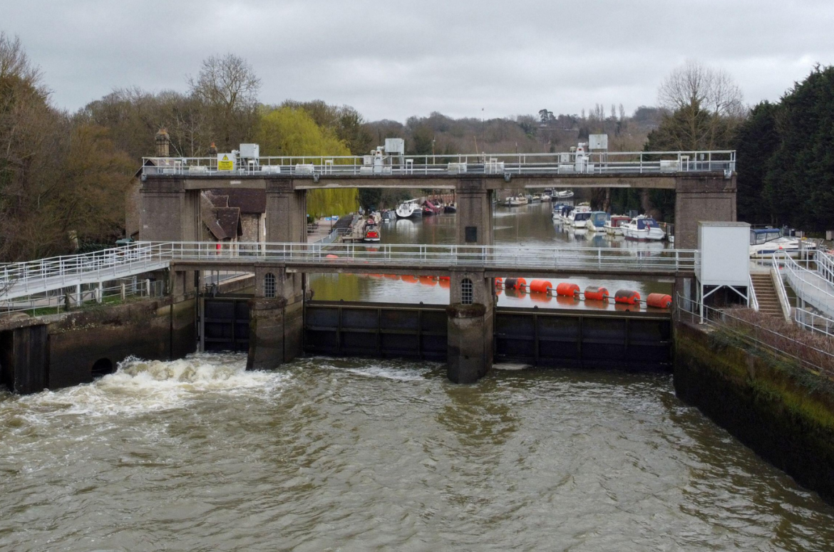 Allington Weir on the River Medway, Kent. Photographed from above showing trees on either side and a grey, cloudy sky. White water is rushing through the sluice gate and boats can be seen on the higher side of the river beyond, behind large orange bouys laid out across the weir.