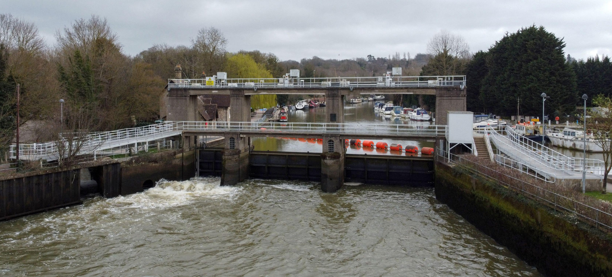 Allington Weir on the River Medway, Kent. Photographed from above showing trees on either side and a grey, cloudy sky. White water is rushing through the sluice gate and boats can be seen on the higher side of the river beyond, behind large orange bouys laid out across the weir.