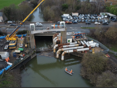 A drone photograph taken above the Woodhouse Mill Regulator. A series of large white pipes run from the riverside into the dark green river water. A yellow crane can be seen on the left of the picture and around 30 cars and vehicles parked nearby to the right of the picture. Workmen in orange hi vis are in a small boat in the river, and on either side of the regulator.