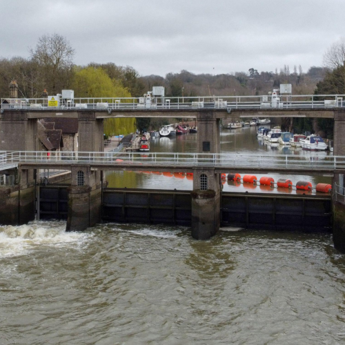 Allington Weir on the River Medway, Kent. Photographed from above showing trees on either side and a grey, cloudy sky. White water is rushing through the sluice gate and boats can be seen on the higher side of the river beyond, behind large orange bouys laid out across the weir.