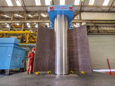 An engineer wearing red overalls stands beside a vertical plate bending machine loaded with a curved metal sheet.