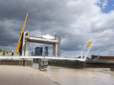Hull's Millennium Bridge photographed from the bank of the River Hull. The bridge is white and forms an 'L' shape with a striking yellow counter-weight on its elevated side. The skiy is blue and full of white/grey clouds. The river is light-brown and the Hull Tidal Barrier can be seen in the distance, behind the bridge.