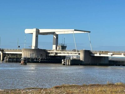 A white, dutch-style road bridge in its closed position to allow traffic to pass over the River yare. Pictured from a distance on a clear, sunny day against a blue sky.