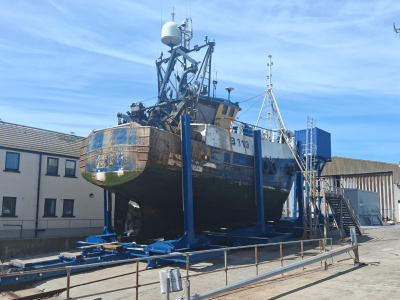 A royal blue painted steel cradle sits at the top of a harbour slipway. A boat is held in the cradle's vertical bilge arms, which stand along the cradle on either side of the boat. The sky is cloudless and blue, and and we can see the grey, concrete slipway in the foreground of the image, a grey, pipework metal fence and cream-coloured buildings with dark rectangular windows at the slide of the slipway.