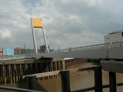 The Millennium Bridge photographed from the side of the river Hull against a blue, cloudy sky. The bridge is 'L' shaped and painted white with a bright yellow counterweight at its top edge.